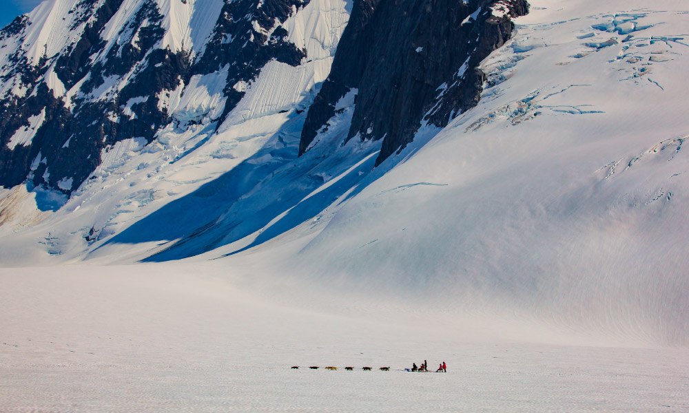 humans on a dogsled look like specks in a massive glacier valley in Alaska